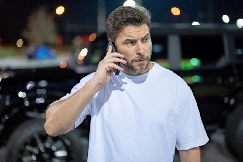 Man talking on a cellphone outdoors at night with a concerned expression near parked cars.