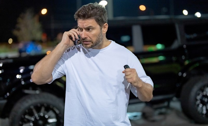 Worried man talking on a cellphone at night while holding car keys near parked vehicles.