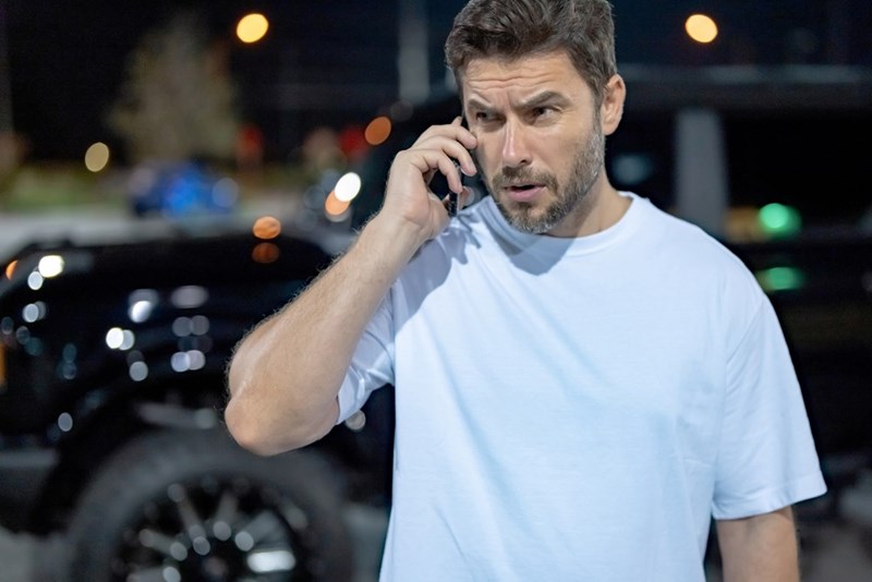 Man talking on a cellphone outdoors at night with blurred city lights and parked vehicles in the background.