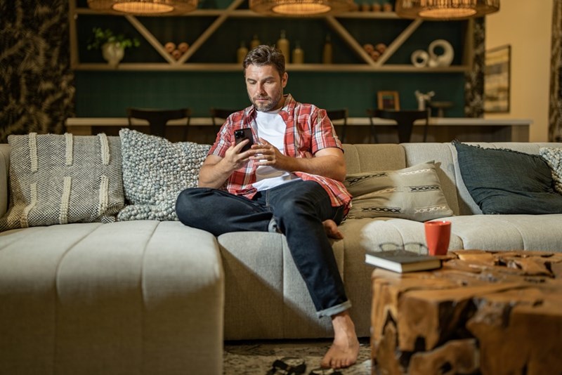 Man sitting on a couch looking at his smartphone in a cozy living room at night.
