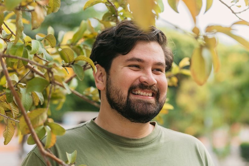 Smiling bearded man standing outdoors beside leafy branches, wearing a green shirt.
