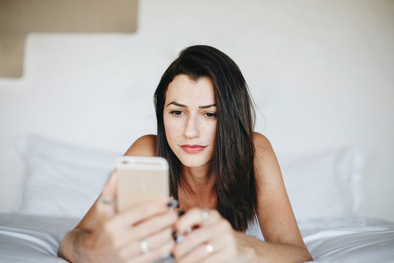 Woman lying on a bed looking at her smartphone with a concerned expression indoors.