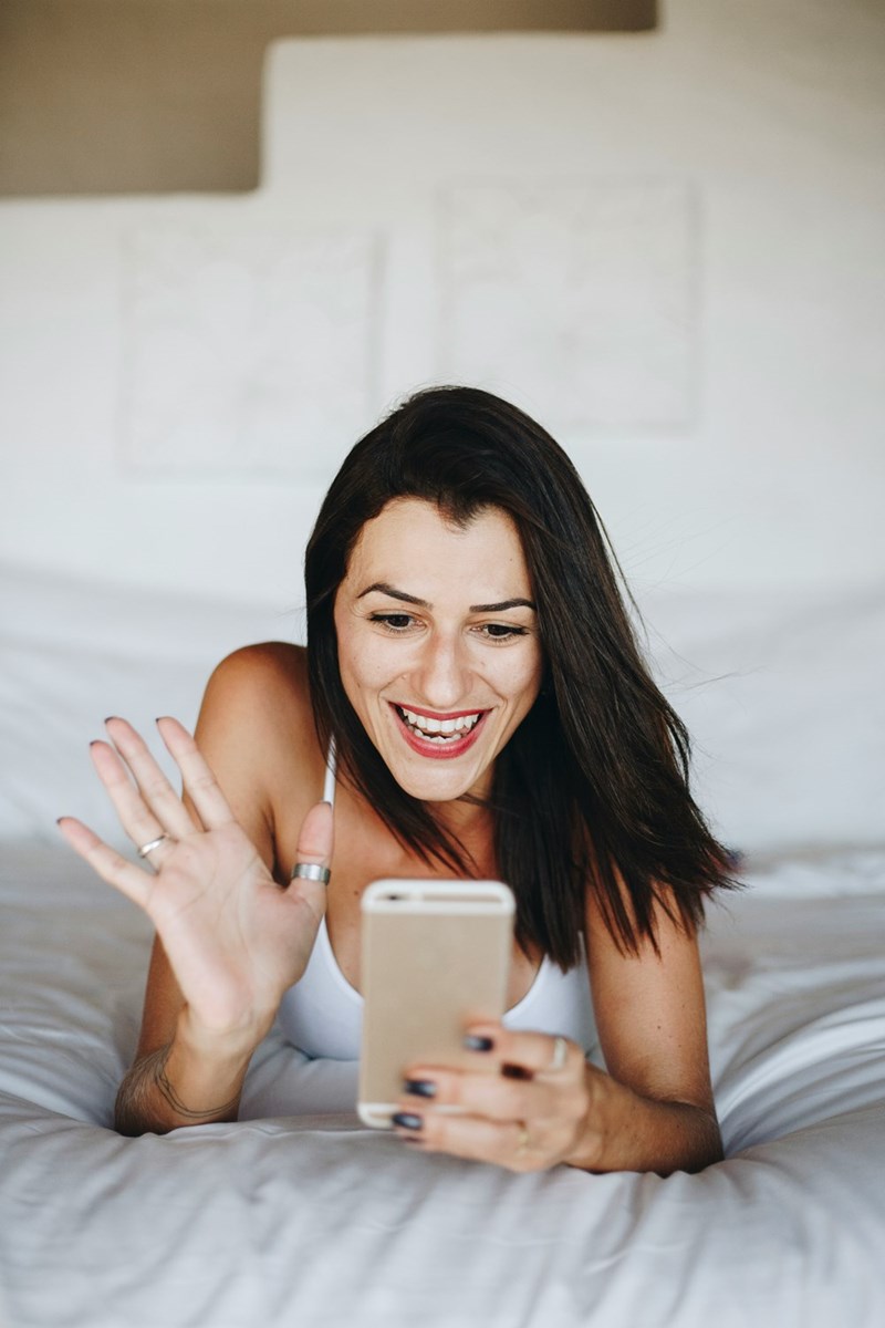 Woman lying on a bed smiling and waving during a video call on her smartphone.
