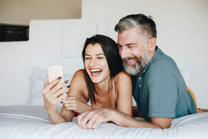Man and woman lying on a bed laughing together while looking at a smartphone.