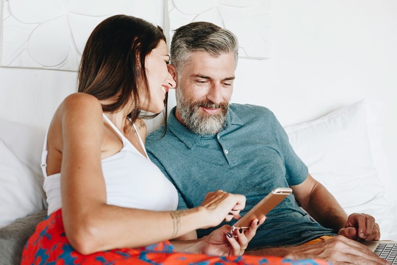 Couple sitting on a bed looking at a smartphone together while using a laptop.