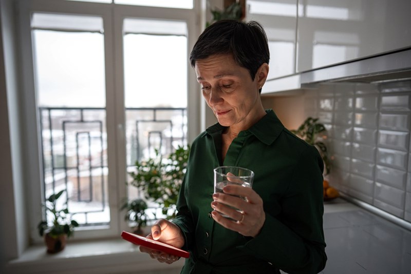 Woman holding a glass of water in a kitchen while checking her smartphone by a bright window.