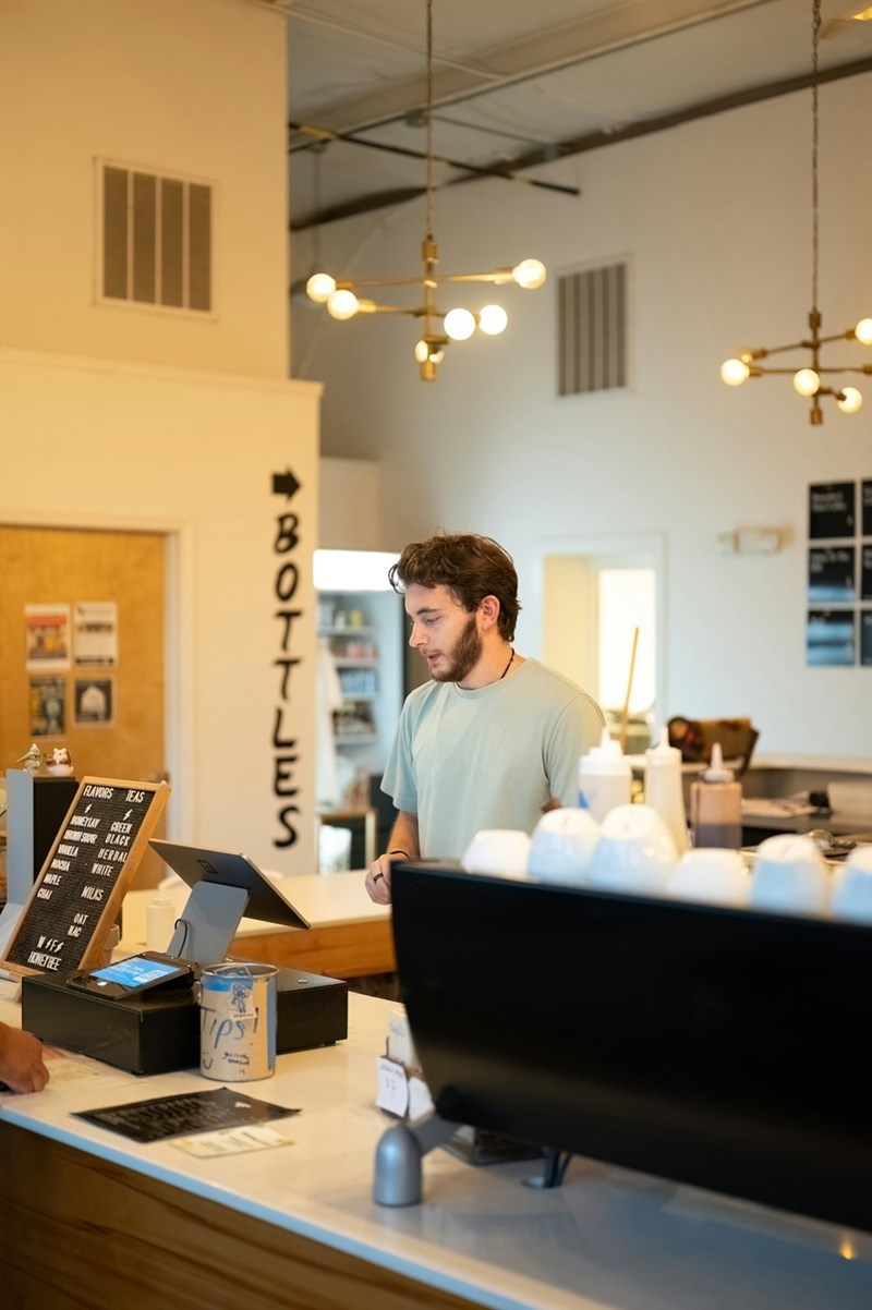 Barista working behind a modern café counter with a tip jar, register, and menu board visible.