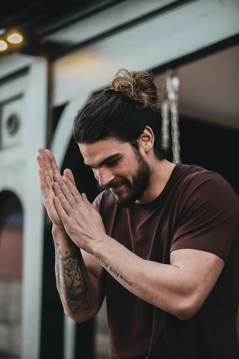 Bearded man with tied-back hair clasping his hands together while standing outdoors.