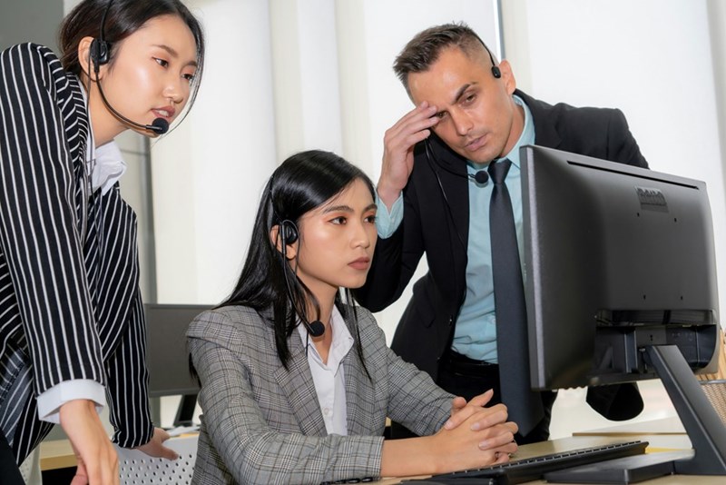 Employees look at computer screen with irritation, while one coworker touches his forehead, as portrayed by models.