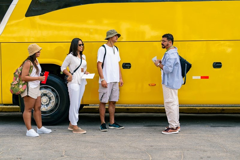 3 tour bus passengers wait outside the bus while a tour guide talks to them.