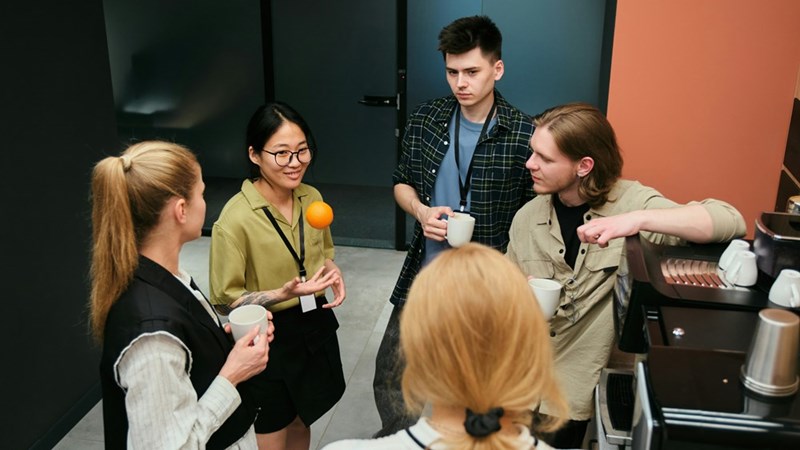 Group of coworkers talk in the office kitchen, trying to figure out why their coworker Sarah has been leaving cryptic lunch notes.