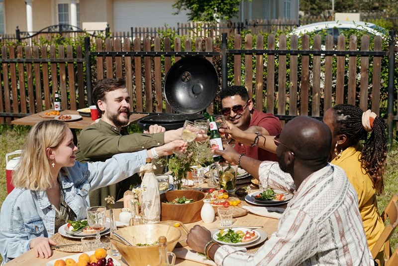 A depiction of five neighborhood residents toasting at an outdoor table behind a built fence.
