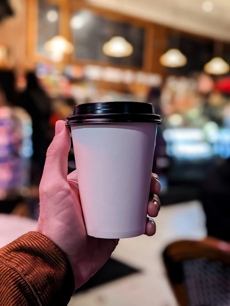 A person holds a to-go coffee cup to take a photograph of with a blurred cafe in the background