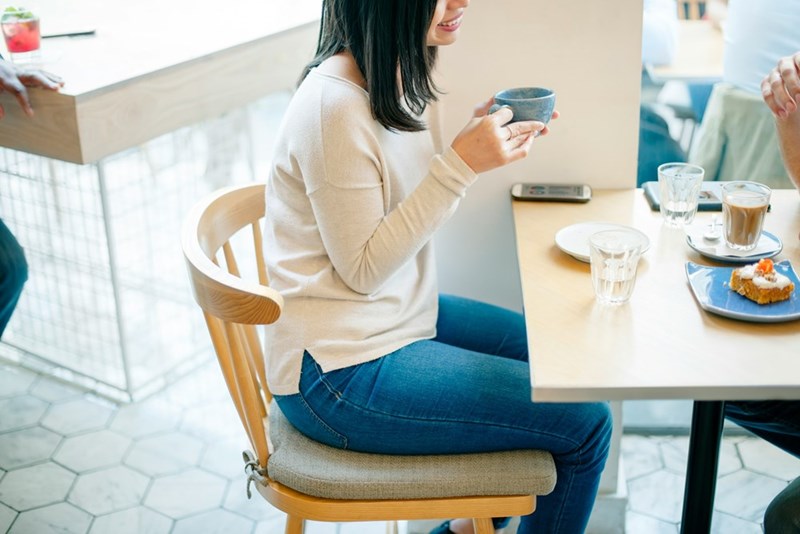 A young woman models sitting at a cafeteria table and sipping on a ceramic mug while enjoying a sweet treat.
