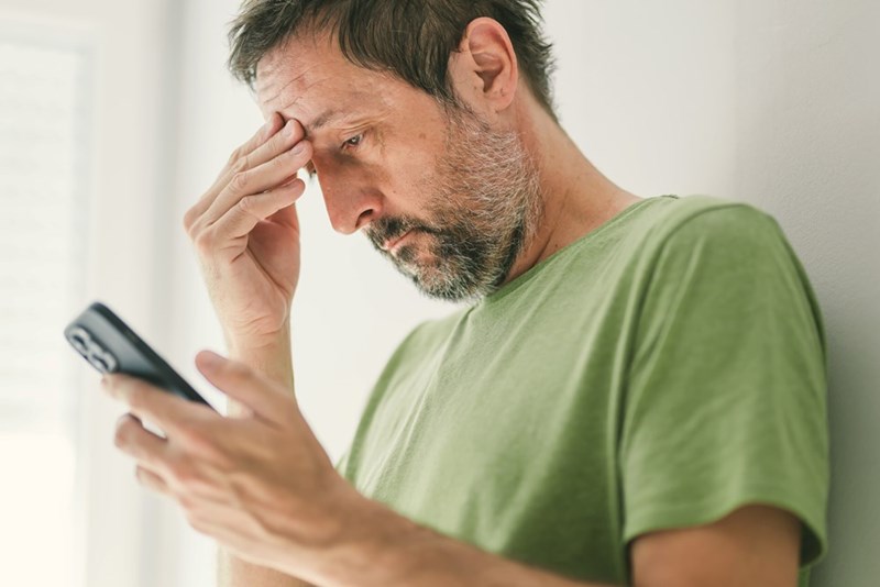 Worried man in a green T-shirt reading his phone while touching his forehead indoors.
