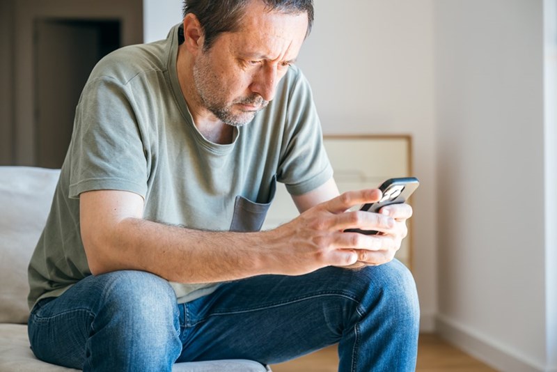 Tense man sitting indoors and looking at his phone with a serious, worried expression.