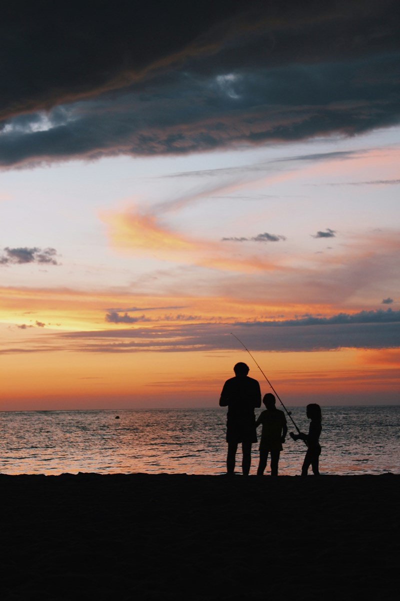 Silhouetted adult and children fishing on a beach at sunset beneath clouds.