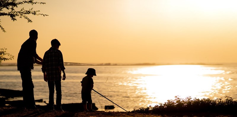 Silhouetted adult and children fishing by the water at sunset with fishing rods.