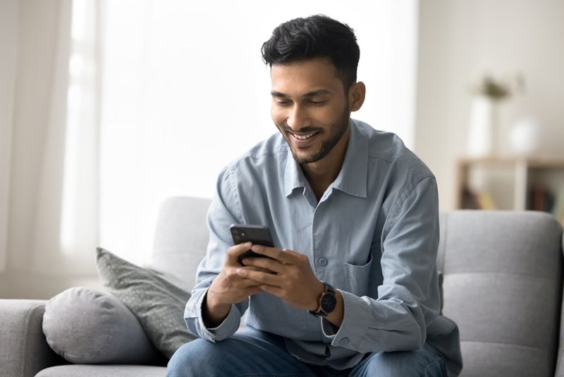 Smiling man sitting on a gray couch and reading his phone in a bright living room.