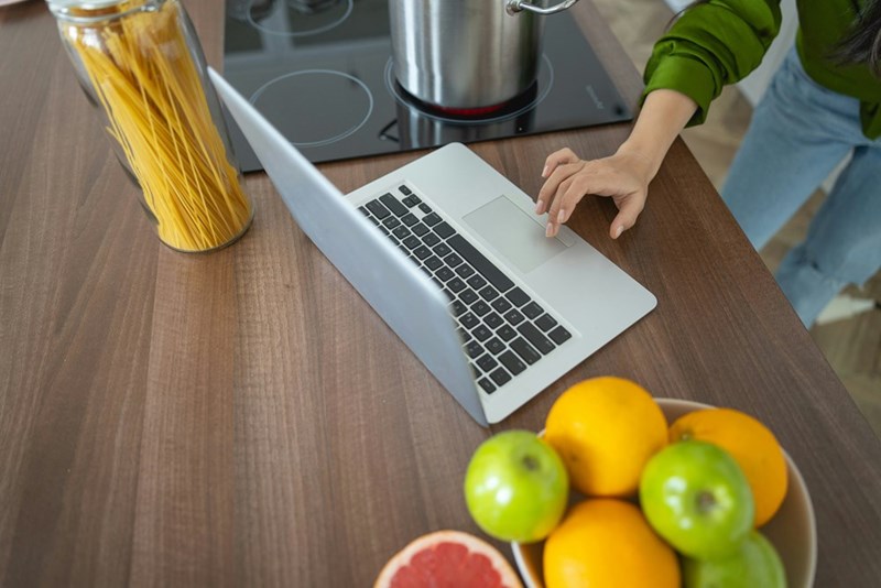 A laptop laying on a kitchen table with fruit and pasta by its side and a hand typing on the keyboard.