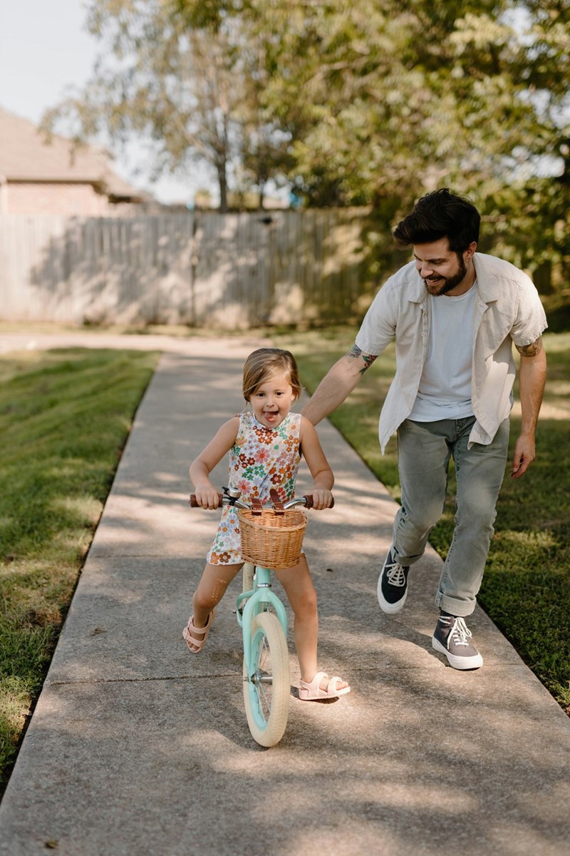 Representation of a father playing with his daughter, helping her to ride her bike outdoors.