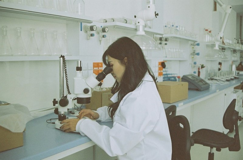 A woman model poses in a white coat while peering into a microscope, appearing to do scientific research.