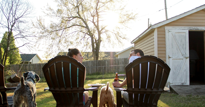 A representation of a male and female couple sitting next to each other in a backyard, turned to each other as if having an argument