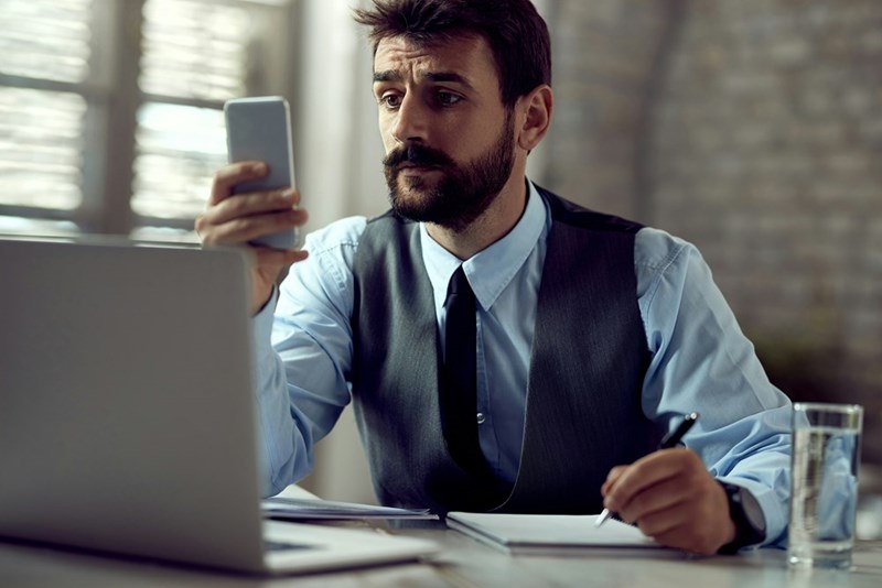 A photo depicting a man in corporate attire looking alarmed at his phone while sitting at his desk with his laptop open.