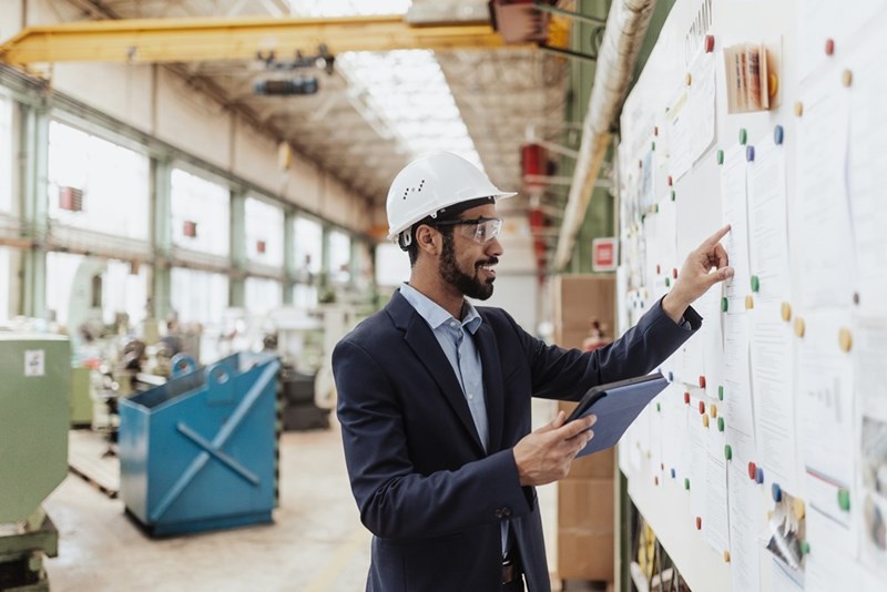 A representation of an operations manager at a manufacturing company wearing a hard hat while looking at a whiteboard.