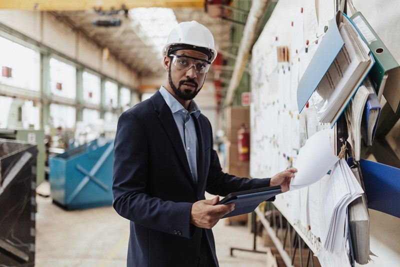 A photograph of a model wearing business attire, looking up from his clipboard in a warehouse environment.