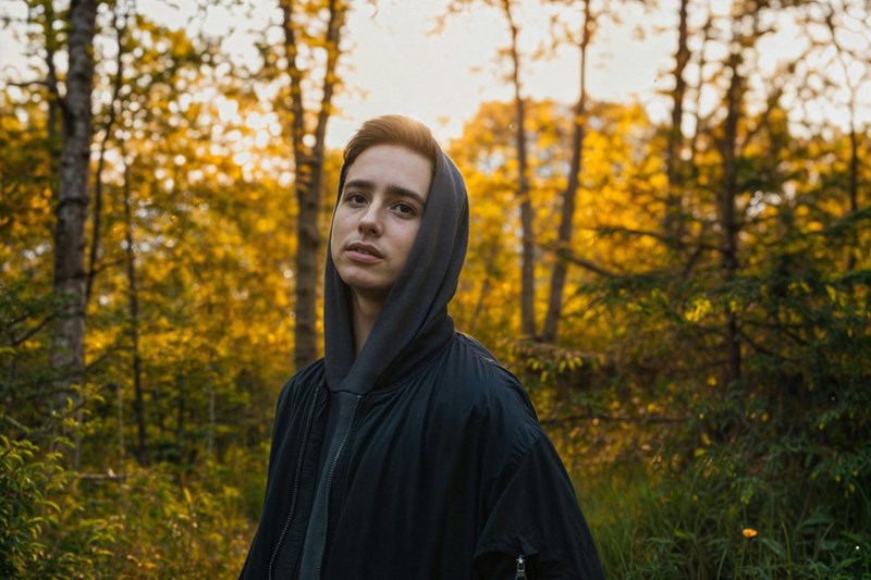 Young man in a dark hoodie standing alone in a sunlit forest with a serious expression.