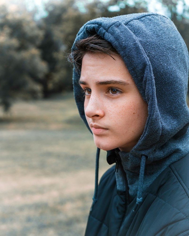 Young man in a dark hoodie looking serious outdoors with trees blurred in the background.