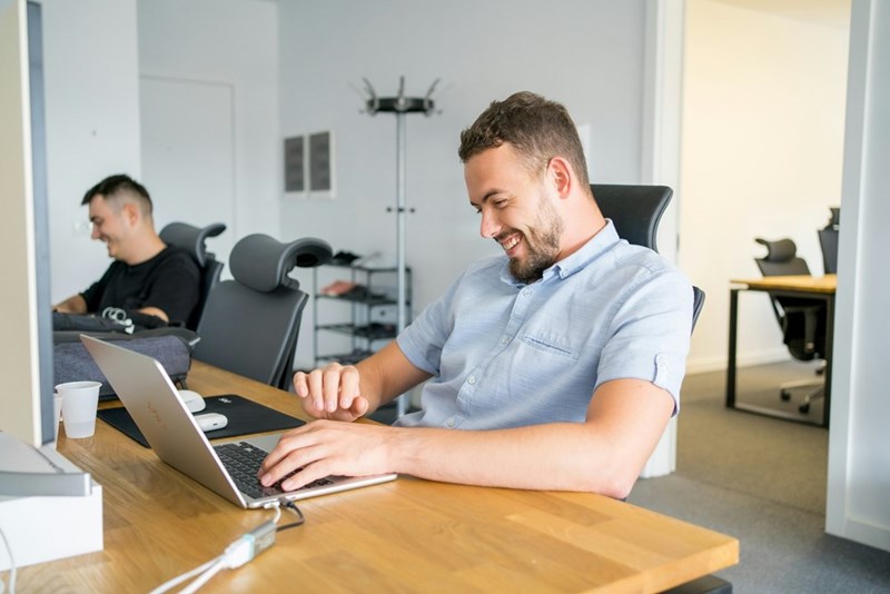 Smiling man working on a laptop at an office desk with another coworker in the background.