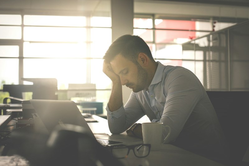 Stressed man leaning over a laptop at an office desk with his hand on his forehead.