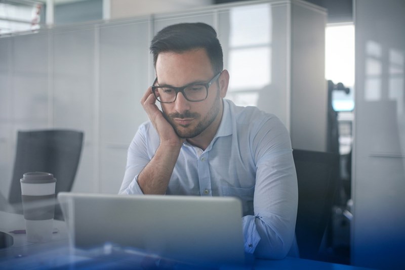 Serious man in glasses sitting at an office desk and looking at a laptop.