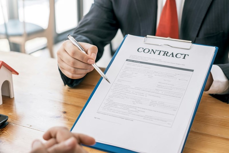 A photograph representing a man in a business suit presenting a contract to someone across a table.