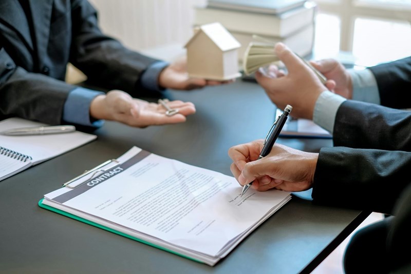 An image depicting a man signing a contract while speaking to a person in corporate attire across a table.