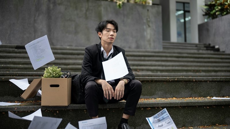 An image of a man (not the actual subject) sitting on the steps of a building in business attire next to a cardboard box of personal items and a series of documents.