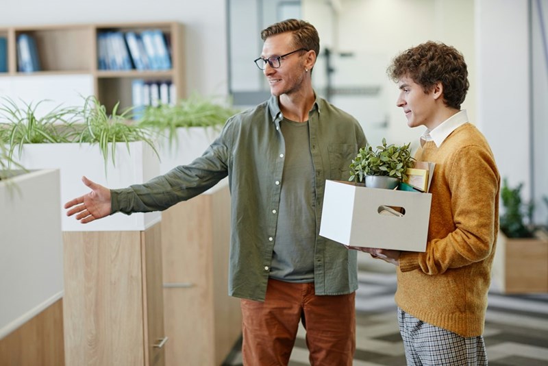 HR manager shows a recently-fired employee to the door while the employee clutches his belongings, as 2 models are demonstrating.