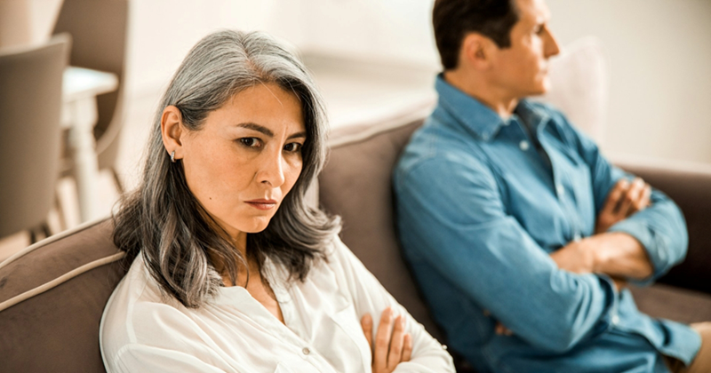 Two models representing a middle-aged male and female couple sitting at opposite ends of a couch with their arms crossed