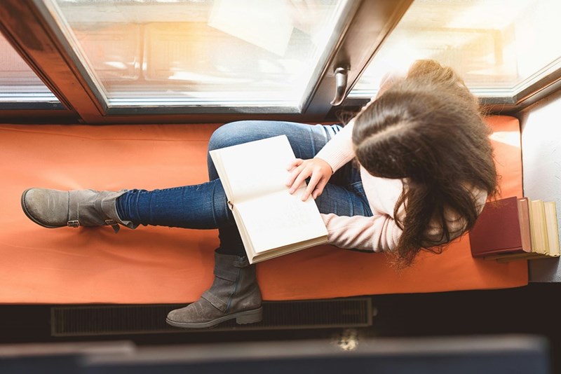Teen girl is depicted by a model attempting to read a book on a comfy bench.