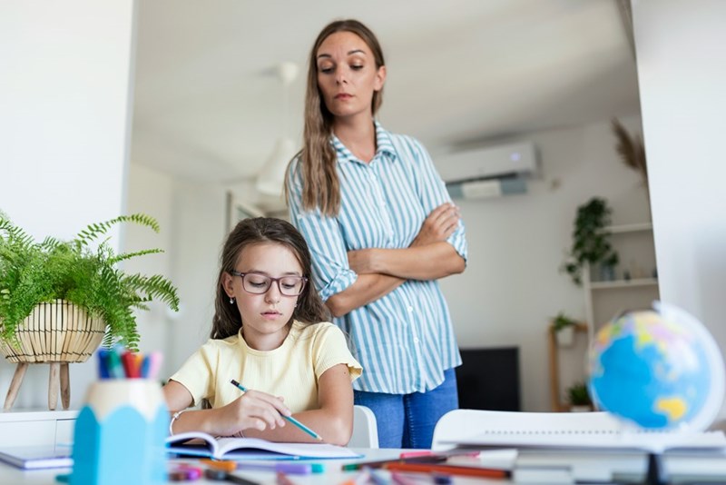 Homeschool Mom peers over her daughter's shoulder as the kid does her schoolwork, in a representation by 2 models.
