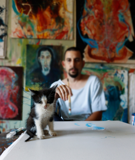 A man paints on a big canvas on the table while a kitten is sitting on the same canvas, both depicted by models.