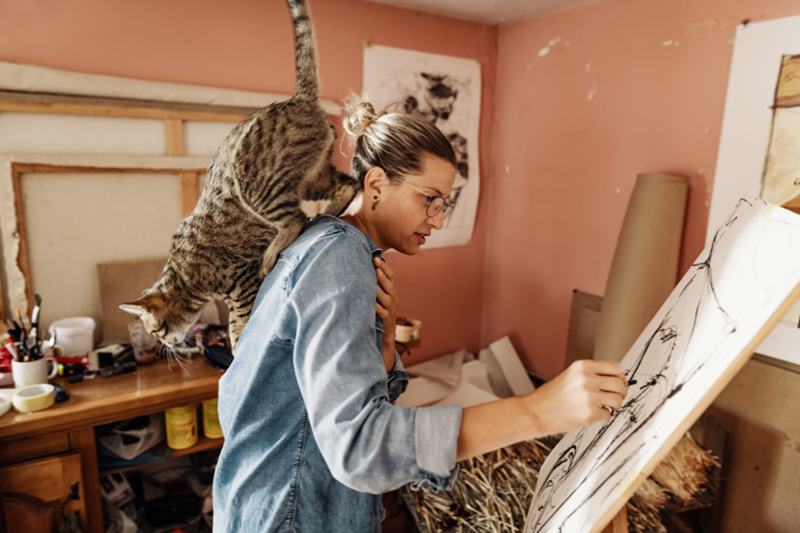 Woman drawing on a canvas while a cat that was sitting on her shoulder jumps off her back, portrayed by human and feline models.