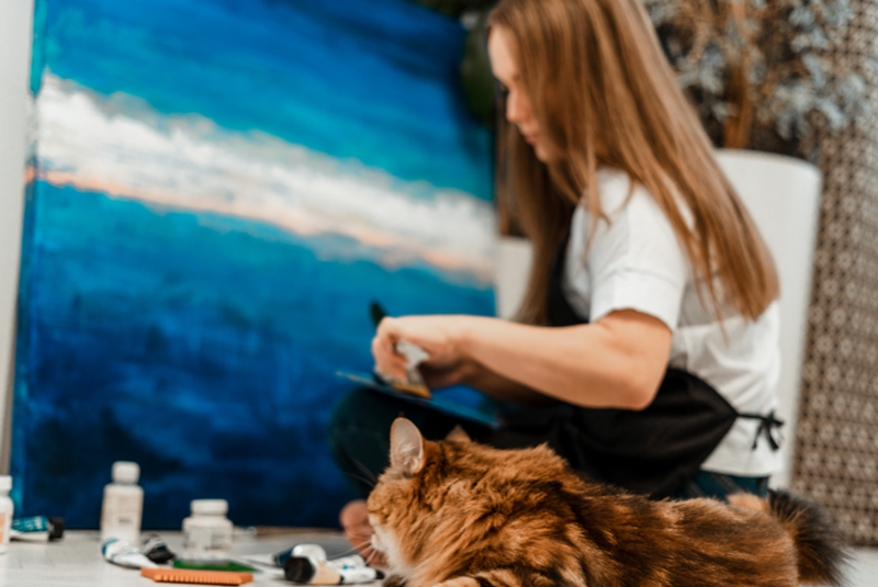 Woman sitting on the floor painting a large canvas, a cat sitting next to her curious about her paint tubes, embodied by a woman model and a cat model.