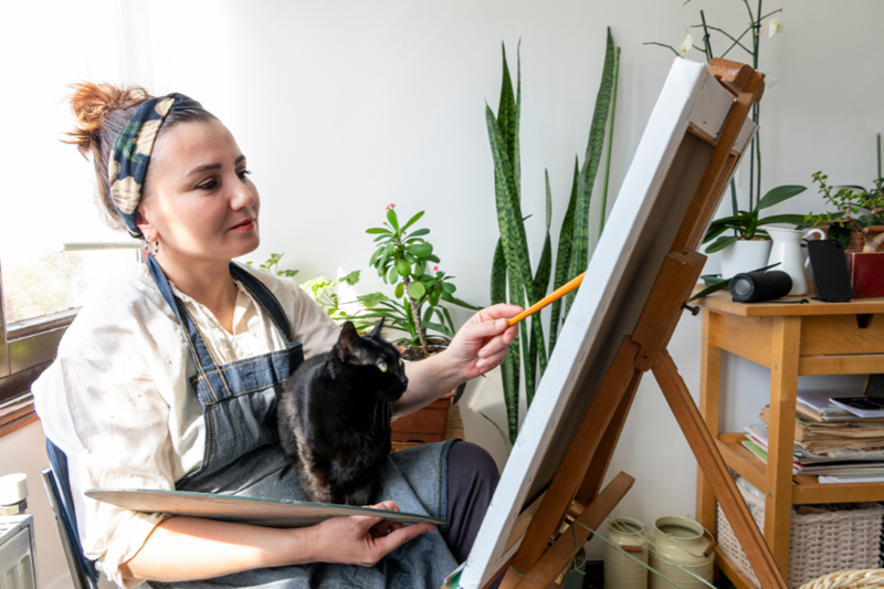 A painter sitting in front of a canvas with a black cat on her lap, the cat looks at the painting, both are models for the picture.