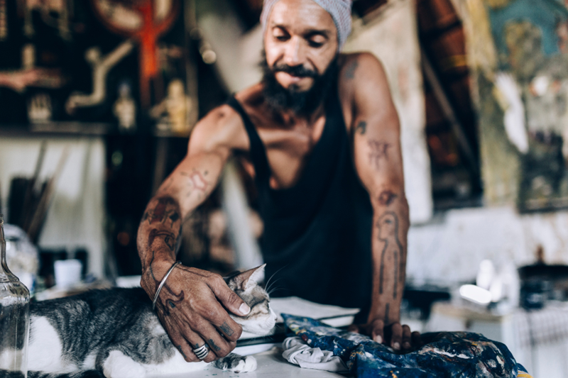 Tattooed artist takes a break from painting in the studio to pet his cat on the table, the two are representing models for the picture.