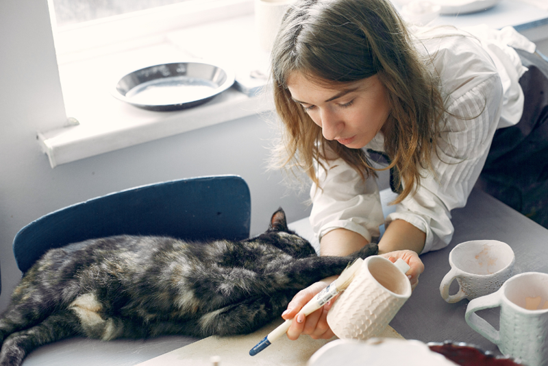 Woman trying to paint mugs but is interrupted by her cat who hugs her hand, acted by human and feline models.