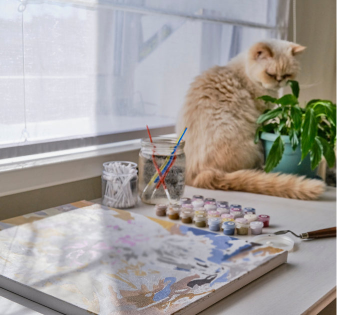 An orange cat is sitting on a table next to a painted canvas, represented by a model cat.