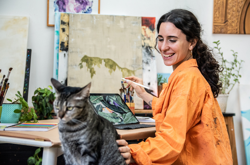 Digital artist stops her work on the iPad to turn around and pet her cat, depicted by woman and cat models.